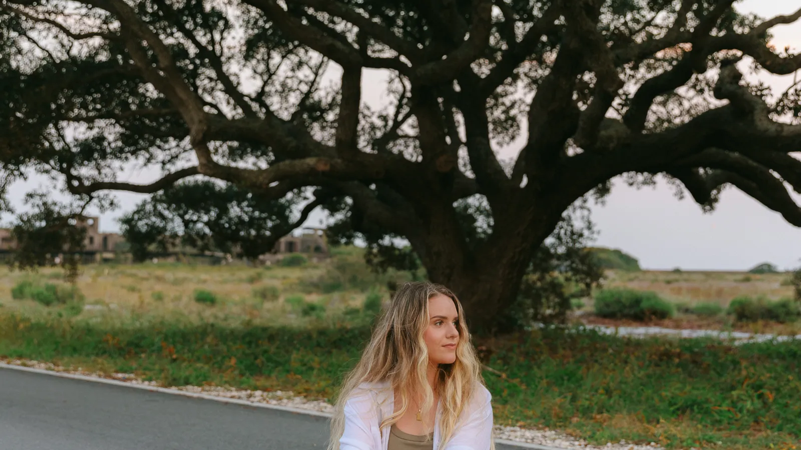 Ali sitting quietly on a sunlit road beneath an oak tree — a moment of stillness